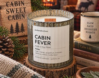 May include: A cylindrical candle in a corrugated metal container with a white label reading Cabin Fever, anchored northwest, made in the PNW, and a list of scent notes including sage, orange, grapefruit, lavender, oakmoss, amber, and tonka bean.