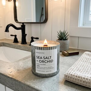 May include: A lit candle in a galvanized metal container with a label that reads "SEA SALT + ORCHID." The candle sits on a concrete countertop next to a small succulent in a gray pot and a box of matches. A white towel is folded nearby.