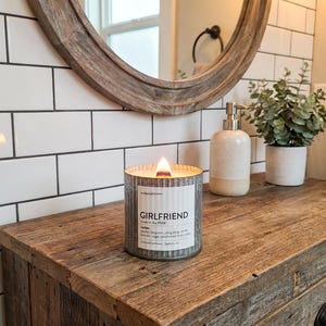 May include: A burning candle in a corrugated metal container, labelled "GIRLFRIEND". The candle rests on a reclaimed wood surface. A round mirror and a small plant are visible in the background. The candle is part of a home fragrance collection.