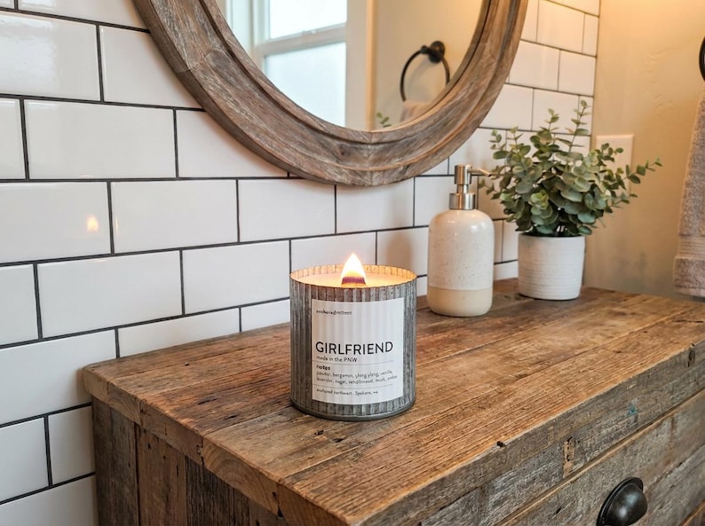May include: A burning candle in a corrugated metal container, labelled "GIRLFRIEND". The candle rests on a reclaimed wood surface. A round mirror and a small plant are visible in the background. The candle is part of a home fragrance collection.
