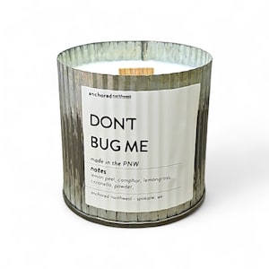 May include: A cylindrical candle in a galvanized metal container. The candle has a wooden wick and a white wax interior. The label reads "DON'T BUG ME" and lists the scent notes. The brand name is "anchored northwest".