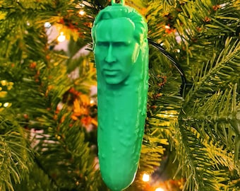 May include: A green pickle-shaped Christmas ornament with a human face. The ornament is hanging on a Christmas tree.