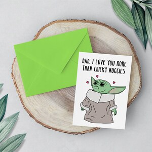 Puede incluir: Una tarjeta verde con un sobre blanco sobre una superficie de madera. La tarjeta presenta un personaje de dibujos animados con el texto "Pap&aacute;, te quiero m&aacute;s que las nugguets de pollo".
