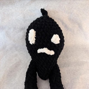 Puede incluir: Juguete de peluche de crochet negro con una cara blanca y una expresión de enfado. El juguete tiene dos brazos pequeños y dos piernas.
