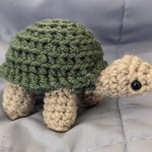 Op de afbeelding: Een gehaakt schildpad speelgoed met een groene schil en een beige lichaam. De schildpad heeft zwarte ogen en een kleine, puntige neus.