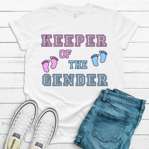 May include: White t-shirt with the text "KEEPER OF THE GENDER" in a varsity style font. The text is outlined in black and has pink and blue baby footprints on either side of the word "THE".