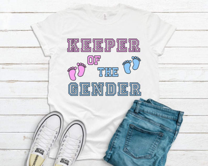 May include: White t-shirt with the text "KEEPER OF THE GENDER" in a varsity style font. The text is outlined in black and has pink and blue baby footprints on either side of the word "THE".