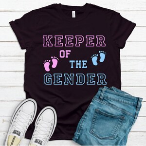 May include: Black t-shirt with the text "KEEPER OF THE GENDER" in white outlined letters. Pink and blue footprints are on either side of the word "THE".