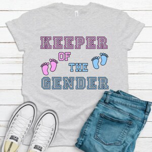 May include: Grey t-shirt with the text "KEEPER OF THE GENDER" in a varsity style font. Pink and blue footprints are above the text.