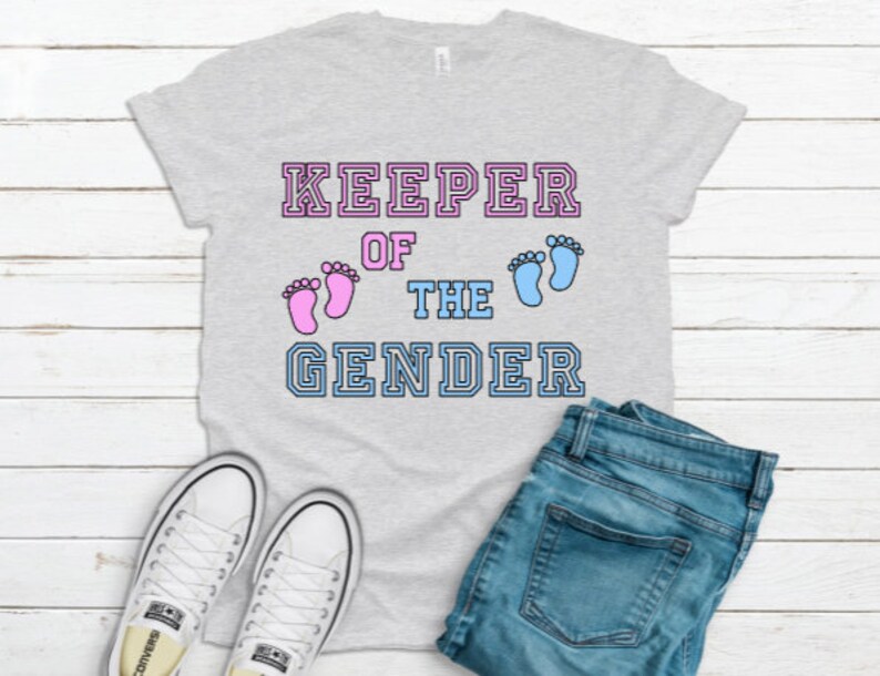 May include: Grey t-shirt with the text "KEEPER OF THE GENDER" in a varsity style font. Pink and blue footprints are above the text.