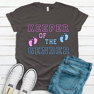 May include: A dark grey t-shirt with the text "KEEPER OF THE GENDER" in white with blue and pink outlines. The text is surrounded by two footprints, one pink and one blue.