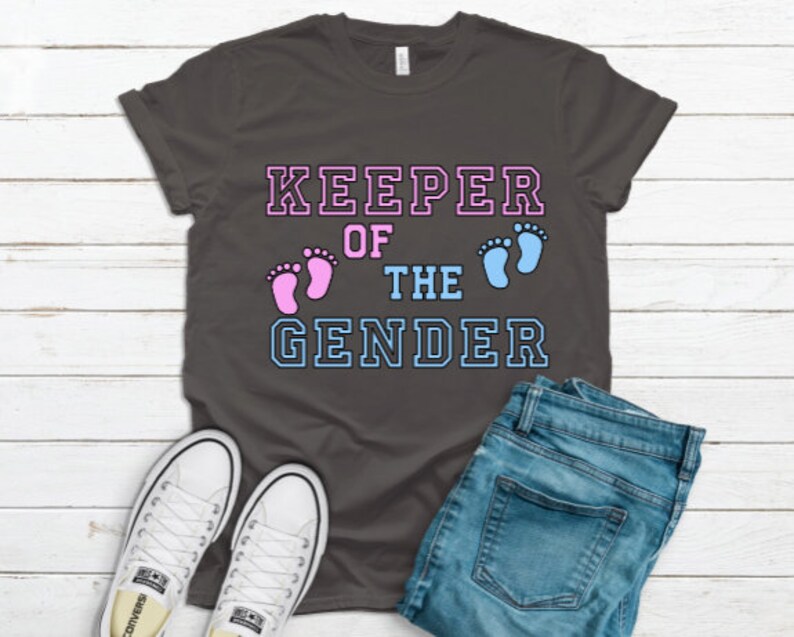 May include: A dark grey t-shirt with the text "KEEPER OF THE GENDER" in white with blue and pink outlines. The text is surrounded by two footprints, one pink and one blue.