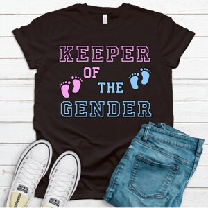 May include: Black t-shirt with the text "KEEPER OF THE GENDER" in white with blue and pink outlines. The text is surrounded by two footprints, one pink and one blue.