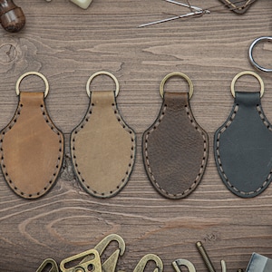 May include: Four leather keychains with stitched edges in brown, tan, dark brown, and black. The keychains are hanging on a wooden surface.