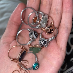 May include: A collection of handmade wire-wrapped rings. The rings feature various gemstones and beads, including a blue-goldstone, a green stone, and a black and white striped bead. The wire is silver and copper-colored.