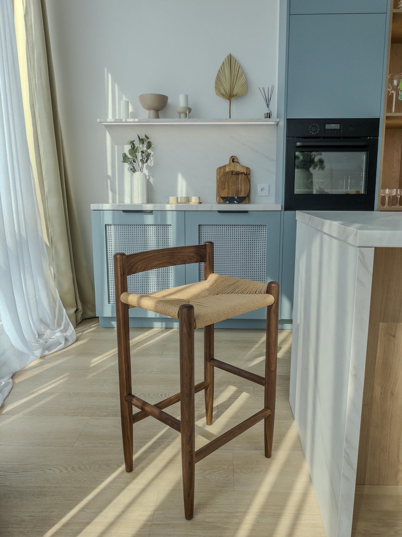 May include: A dark brown wooden bar stool with a woven cane seat. The stool is in a modern kitchen with white walls and blue cabinets.