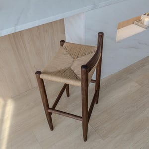 May include: A dark brown wooden stool with a woven straw seat. The stool is in a kitchen setting with a white countertop and light wood floor.