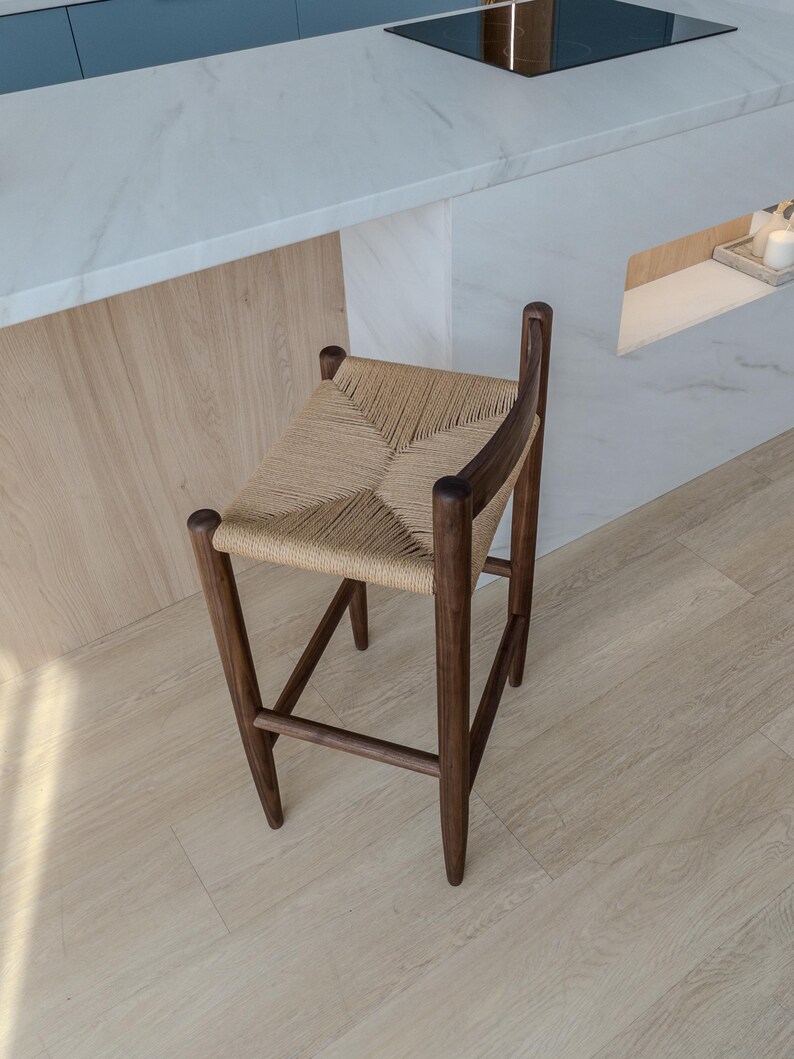 May include: A dark brown wooden stool with a woven straw seat. The stool is in a kitchen setting with a white countertop and light wood floor.