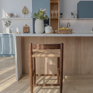 May include: A wooden bar stool with a woven cane seat. The stool is brown and has a natural finish. It is sitting on a light wood floor.