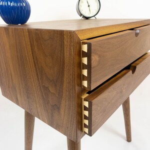 May include: A wooden nightstand with two drawers and tapered legs. The drawers have a dovetail joint detail and are made of walnut wood.