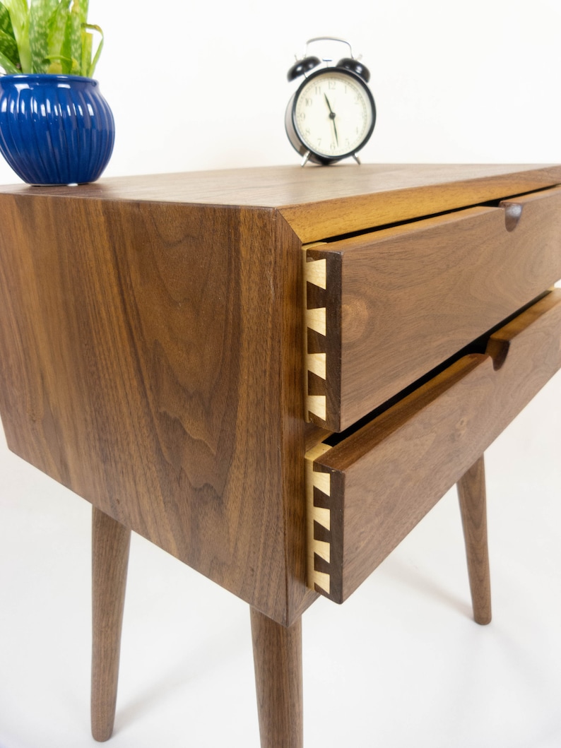 May include: A wooden nightstand with two drawers and tapered legs. The drawers have a dovetail joint detail and are made of walnut wood.