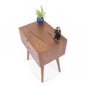 May include: A wooden end table with two drawers and four legs. The table is made of walnut wood and has a natural finish. A small blue planter with a green plant is on the table, along with a black alarm clock.