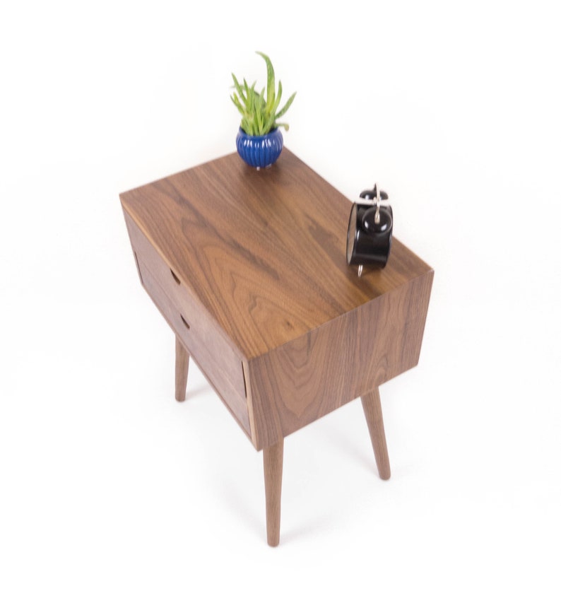 May include: A wooden end table with two drawers and four legs. The table is made of walnut wood and has a natural finish. A small blue planter with a green plant is on the table, along with a black alarm clock.