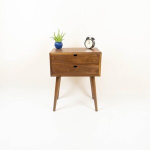 May include: A wooden nightstand with two drawers and tapered legs. A small potted plant and a black alarm clock are on top of the nightstand.
