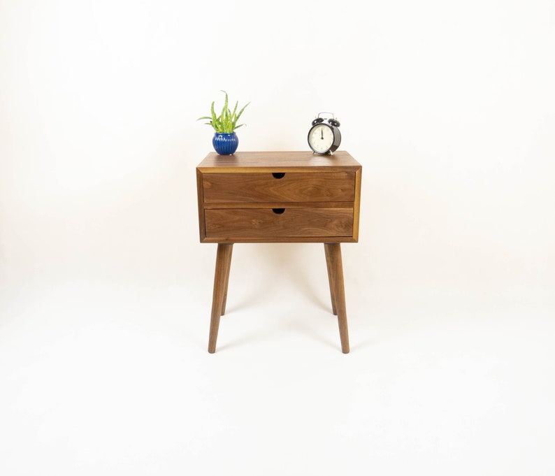 May include: A wooden nightstand with two drawers and tapered legs. A small potted plant and a black alarm clock are on top of the nightstand.