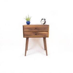 May include: A two-drawer wooden nightstand with a dark walnut finish. The nightstand has tapered legs and black metal drawer pulls. A small potted plant and a black alarm clock are on top of the nightstand.