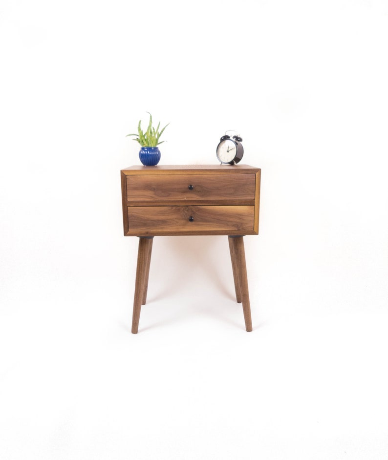 May include: A two-drawer wooden nightstand with a dark walnut finish. The nightstand has tapered legs and black metal drawer pulls. A small potted plant and a black alarm clock are on top of the nightstand.