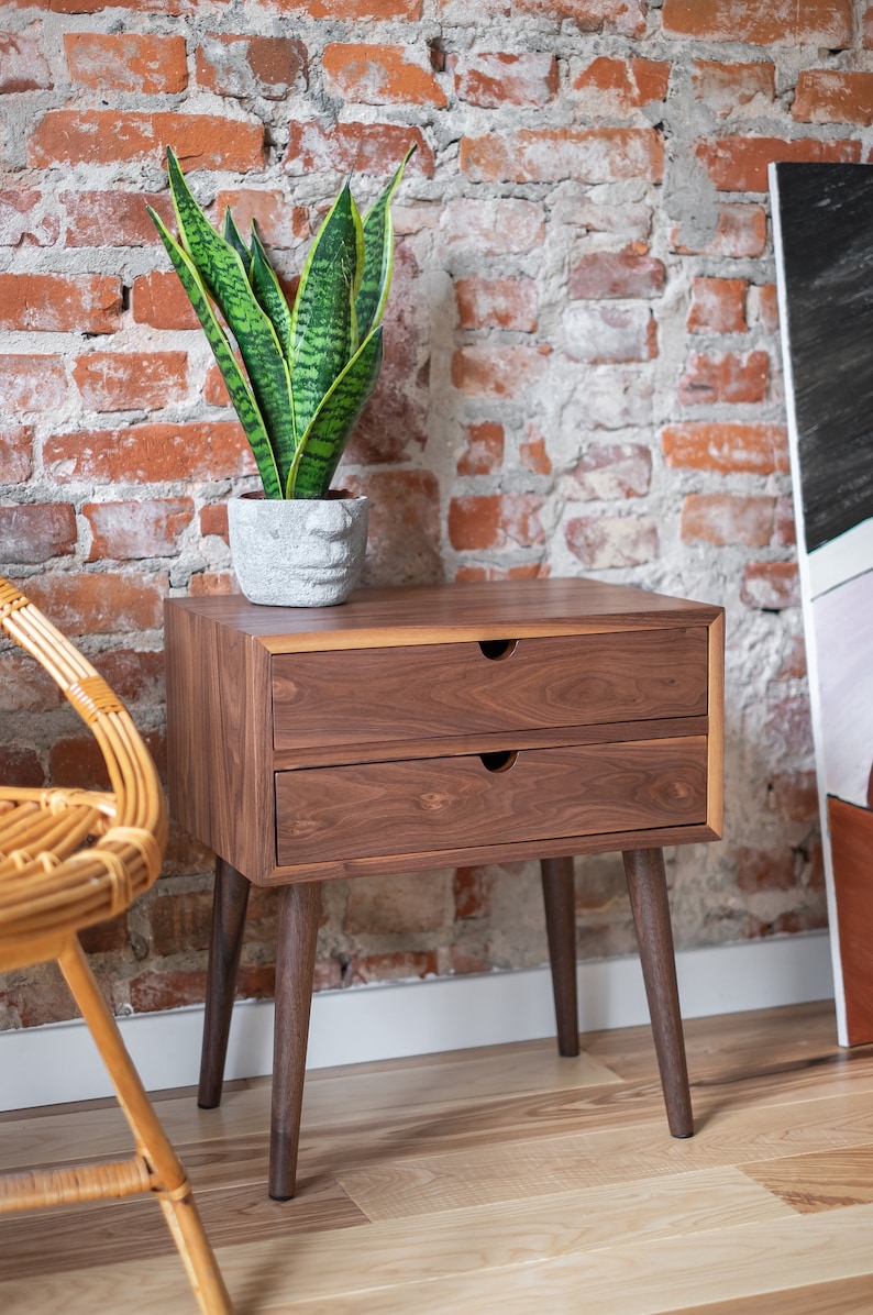 May include: A dark brown wooden nightstand with two drawers and tapered legs. The nightstand is in front of a brick wall and a potted plant.