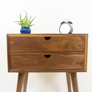 May include: A two-drawer wooden nightstand with a walnut finish. The nightstand has a simple design with rounded legs and black drawer pulls. A small potted plant and a black alarm clock are on top of the nightstand.