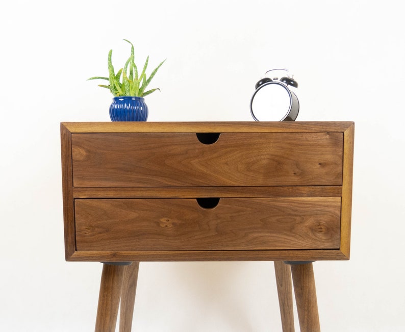 May include: A two-drawer wooden nightstand with a walnut finish. The nightstand has a simple design with rounded legs and black drawer pulls. A small potted plant and a black alarm clock are on top of the nightstand.