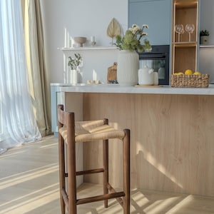 May include: A wooden bar stool with a woven rattan seat. The stool is brown and has a simple, modern design. It is sitting in a kitchen with a white countertop and a light wood backsplash.