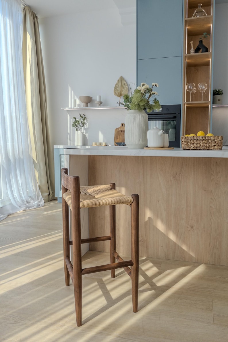 May include: A wooden bar stool with a woven rattan seat. The stool is brown and has a simple, modern design. It is sitting in a kitchen with a white countertop and a light wood backsplash.