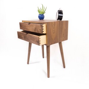 May include: A wooden nightstand with two drawers and four legs. The drawers are open and the wood grain is visible. A small potted plant and an alarm clock are on top of the nightstand.