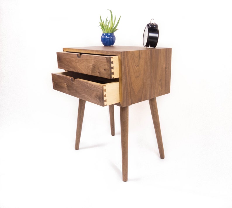 May include: A wooden nightstand with two drawers and four legs. The drawers are open and the wood grain is visible. A small potted plant and an alarm clock are on top of the nightstand.