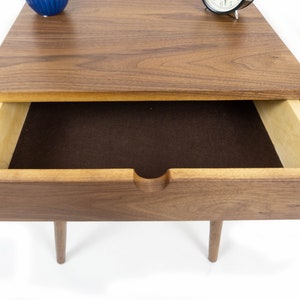 May include: A wooden nightstand with a single drawer. The drawer is open and lined with brown felt. The nightstand has a walnut finish and is sitting on a white surface.