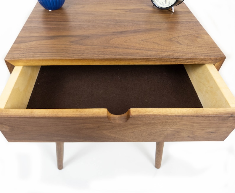 May include: A wooden nightstand with a single drawer. The drawer is open and lined with brown felt. The nightstand has a walnut finish and is sitting on a white surface.
