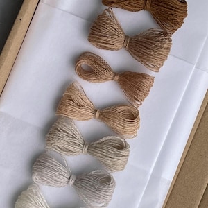 Op de afbeelding: Een verzameling handgemaakte strikken van touw in verschillende bruine en beige tinten, gerangschikt op wit papier. De strikken zijn netjes geknoopt en gepresenteerd in een kartonnen doos, wat een rustieke en natuurlijke uitstraling geeft. Perfect voor knutselprojecten.