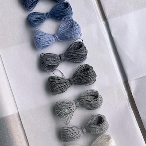 Op de afbeelding: Een assortiment handgemaakte garen strikken in tinten blauw, grijs en wit. De strikken zijn in een rij gerangschikt op een wit oppervlak, waarbij de verschillende kleuren en texturen van het garen worden getoond. Ideaal voor knutselprojecten.