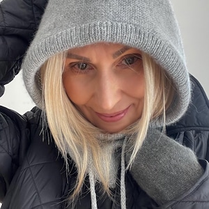 Puede incluir: Una capucha de punto gris con cordón, usada sobre un cuello alto y guantes a juego. La persona lleva una chaqueta acolchada negra. La capucha está hecha de un material de punto suave y texturizado.