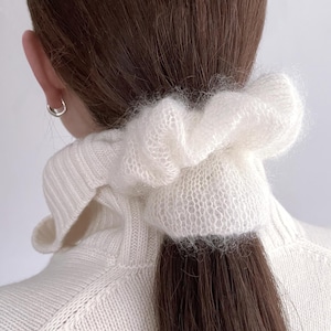 Könnte beinhalten: Ein weißes, flauschiges Scrunchie aus weichem Strickmaterial. Das Scrunchie wird in einem Pferdeschwanz getragen und ergänzt das braune Haar. Die Person trägt einen weißen Pullover. Ein silberner Ohrring ist sichtbar.