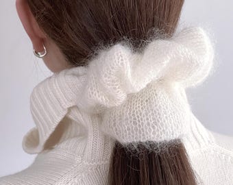 May include: A white, fluffy scrunchie made of a soft, knitted material. The scrunchie is worn in a ponytail, complementing the brown hair. The person is wearing a white sweater. A silver earring is visible.