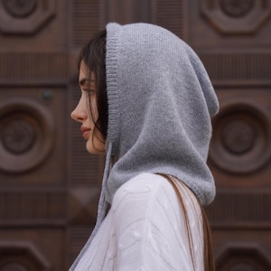 Könnte beinhalten: Eine graue Strickhaube mit Kordelzügen. Die Haube besteht aus einem weichen, strukturierten Material und wird über dem Kopf getragen. Die Haube wird vor einem verschwommenen Holzhintergrund gezeigt. Die Haube ist Teil einer Winterbekleidungskollektion.