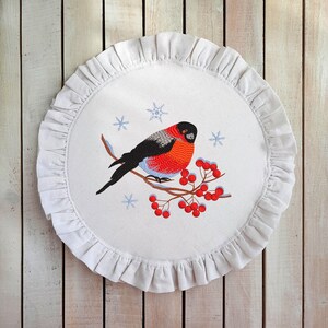Pu&ograve; includere: Un sottobicchiere rotondo in tessuto bianco con un bordo arricciato presenta un disegno ricamato di un pettirosso appollaiato su un ramo con bacche rosse e fiocchi di neve.