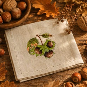 Puede incluir: Una servilleta de lino beige con un diseño bordado de avellanas y hojas. El bordado presenta tonos de verde, marrón y naranja. La servilleta está doblada y colocada sobre una superficie de madera, rodeada de nueces y hojas de otoño.