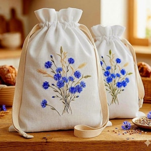 Peut inclure: Deux sacs blancs à cordon de serrage brodés de bleuets bleus et d'épis de blé. Les sacs ont des sangles beiges et sont faits d'un tissu texturé. Ils sont posés sur une surface en bois.