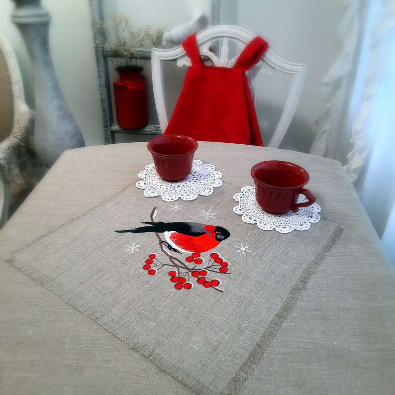 Pu&ograve; includere: Un set di due tazze in ceramica rossa con sottobicchieri in crochet bianco, su un tovaglietta di lino con un ricamo di un pettirosso appollaiato su un ramo con bacche rosse e fiocchi di neve.